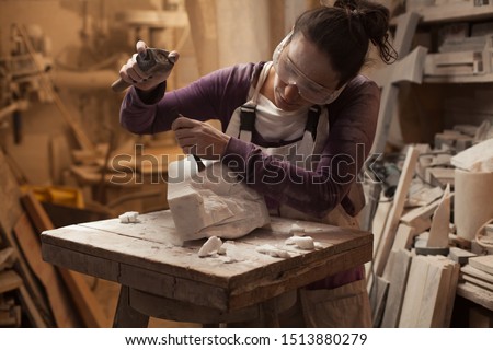 Female sculptor at work in a workshop, using hammer and chisel to sculpt a piece of white marble stone, stonemasonry and stonecraft