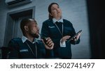 Female security officer stands and uses tablet computer. African American male dispatcher talks in walkie talkie. Multiethnic surveillance operators control CCTV cameras in modern monitoring center.