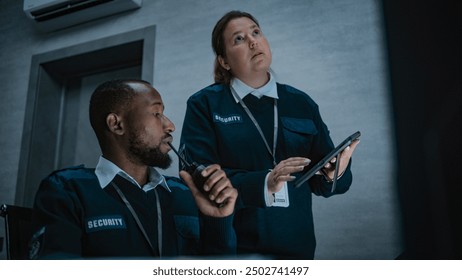 Female security officer stands and uses tablet computer. African American male dispatcher talks in walkie talkie. Multiethnic surveillance operators control CCTV cameras in modern monitoring center. - Powered by Shutterstock