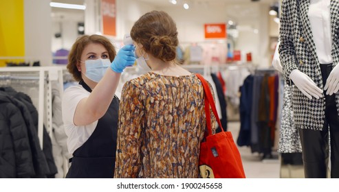 Female Seller In Safety Mask And Gloves Checking Temperature Of Customer Before Entering Clothing Store. Woman Employee Scanning Clients Temperature With Infrared Thermometer In Shopping Mall