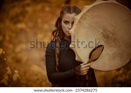 A female shaman in black garment with ethnic ornaments on her face performs a ritual with a shamanic drum in an autumn forest. Ethnic culture. Shamanic rituals. Copy space.