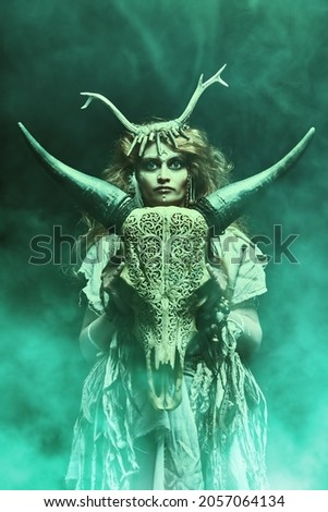 Female shaman in an ethnic dress doing a mysterious ritual with a help of an animal skull. Dark mystical background with green haze. Black magic concept, fantasy. Paganism. Halloween.