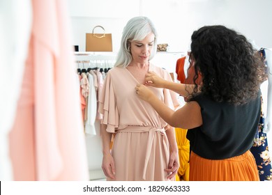 Female shop seller helping customer to try on new dress. Woman choosing clothes in fashion store. Buying clothes in boutique concept - Powered by Shutterstock