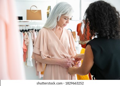 Female shop seller helping woman to try on new dress and tying waistband. Customer choosing clothes in fashion store. Buying clothes in boutique concept - Powered by Shutterstock