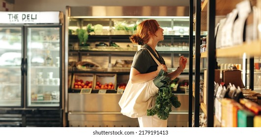 Female Shopper Choosing Food Products From A Shelf While Carrying A Bag With Vegetables In A Grocery Store. Young Woman Doing Some Grocery Shopping In A Trendy Supermarket.