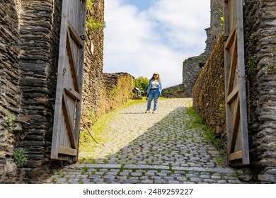 Female sightseer standing in front of main gate of castle ruins of La Roche-en-Ardenne, cobblestone path, stone walls worn by passage of time, casual clothing, sunny day in Belgium - Powered by Shutterstock