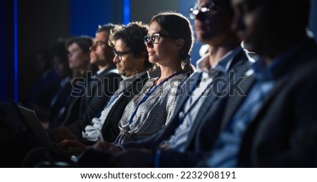Female Sitting in a Dark Crowded Auditorium at a Tech Conference. Young Woman Using Laptop Computer. Specialist Watching Innovative Technology Presentation About New Software and High Tech Products.