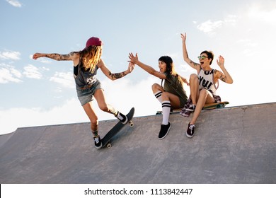 Female skateboarder riding skateboard at skate park with friends sitting on ramp having fun. Woman skater giving high five to female friend sitting on ramp during her routine. - Powered by Shutterstock