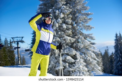 Female Skier In Goggles Shielding From Sun With Her Hand And Looking For Someone. Smiling Woman Coming Up On Slope Top With Ski Lift. Skier Before Freeride-descending. Mountain View On Background.