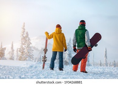 Female skier and male snowboarder are standing on background of snowcapped mountain at sunset time. Ski resort concept