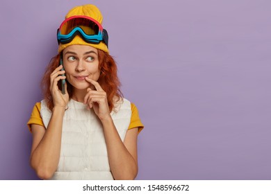 Female Skiier With Ginger Hair Makes Phone Call, Looks Aside With Pensive Expression, Receives Good Suggestion, Enjoys Hobby During Pastime, Isolated On Purple Wall, Phones Friend. Relax Moment