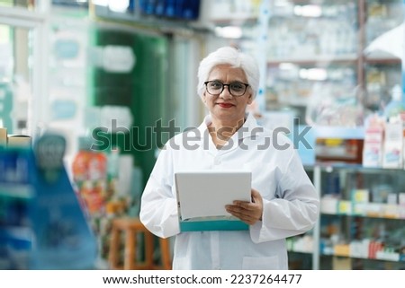 Female smart senior pharmacist in professional white gown looking at camera and checking stock inventory in modern pharmacy, drugstore indoor holding cli. Pharmacy, medicine, and healthcare concept. 
