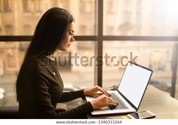 Female smart university student learning online via pc\
laptop computer with empty mock up copy space display background\
for advertising text message, sitting in modern interior near\
window in evening 