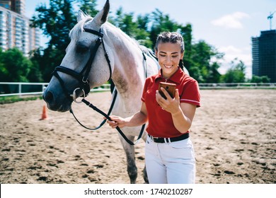 Female Smiling Jockey In Casual Outfit Surfing Internet On Cellphone While Walking Grey Horse On Riding Arena In Summer Afternoon