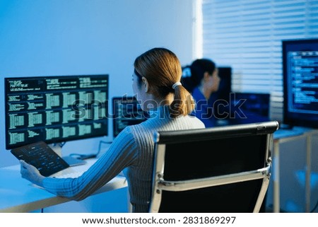Female software developer using a stylus on a digital tablet while analyzing code on multiple computer monitors in a modern tech office.