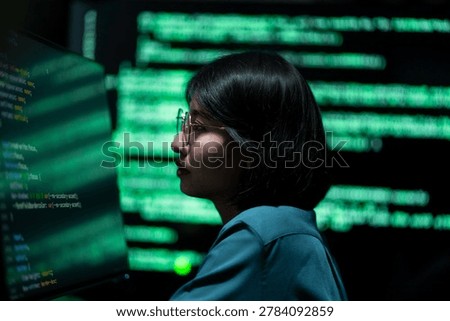 A female software engineer working with digital programming code and cybersecurity data displayed on computer screens in a modern office environment. The scene represents information technology, software development, and data security operations.