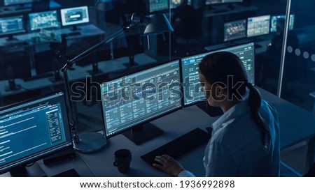 Female Software Engineer Working in a Modern Monitoring Office with Live Analysis Feed with Charts on a Big Digital Screen. Monitoring Room Big Data Scientists and Managers Sit in Front of Computers.