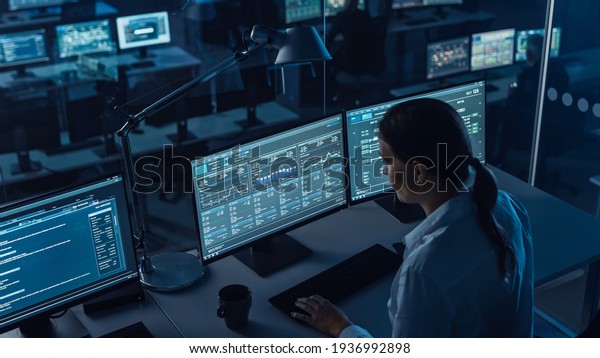 Female Software Engineer Working in a Modern
Monitoring Office with Live Analysis Feed with Charts on a Big
Digital Screen. Monitoring Room Big Data Scientists and Managers
Sit in Front of
Computers.