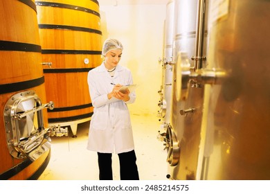 Female sommelier wearing uniform wine expert taking notes collecting wine samples in oak barrels and stainless steel tanks wine fermentation room production factory quality production process.  - Powered by Shutterstock