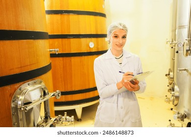 Female sommelier wearing uniform wine expert taking notes collecting wine samples in oak barrels and stainless steel tanks wine fermentation room production factory quality production process.  - Powered by Shutterstock