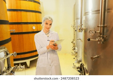 Female sommelier wearing uniform wine expert taking notes collecting wine samples in oak barrels and stainless steel tanks wine fermentation room production factory quality production process.  - Powered by Shutterstock