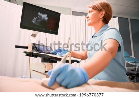 Female sonographer examining woman with ultrasound scanner. Doctor moving ultrasound transducer on patient abdomen while looking at display. Concept of medical examination and ultrasound diagnostics.