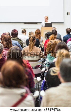 Female speaker giving presentation in lecture hall at university workshop. Audience in conference hall. Rear view of unrecognized participant in audience. Scientific conference event.