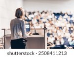 Female speaker giving a talk on corporate business conference. Unrecognizable people in audience at conference hall. Business and Entrepreneurship event