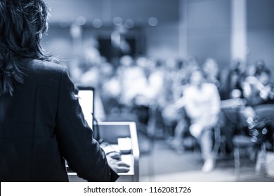 Female Speaker Giving A Talk On Corporate Business Conference. Unrecognizable People In Audience At Conference Hall. Business And Entrepreneurship Event. Black And White, Blue Toned Image.