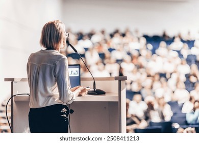 Female speaker giving a talk on corporate business conference. Unrecognizable people in audience at conference hall. Business and Entrepreneurship event