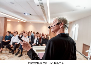 Female speaker in suit with headset and laser pointer looking at screen while giving a talk on corporate business meeting. Audience listening at conference hall. Business and Entrepreneurship event
