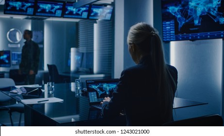 Female Special Agent Works On A Laptop In The Background Special Agent In Charge Talks To A Military Man In The Monitoring Room. Background Busy System Control Center With Monitors Showing Data Flow.