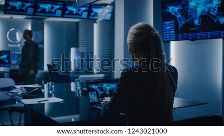 Female Special Agent Works on a Laptop in the Background Special Agent in Charge Talks To a Military Man in the Monitoring Room. Background Busy System Control Center with Monitors Showing Data Flow.