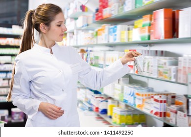 Female specialist is attentively looking medicines near shelves in pharmacy