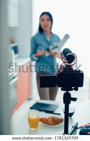 Female specialist having a camera with microphone on a tripod and gesturing while being recorded on a video