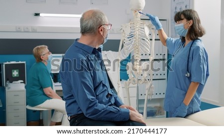 Female specialist pointing at spinal cord on human skeleton, explaining bones anatomy system to old patient at consultation. Assistant talking about osteopathy diagnosis, orthopedic spine.