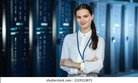 Female IT Specialist is standing at the Camera in Data Center Next to Server Racks and Looking at the Camera. Her Arms Crossed and She Smiles to the Camera.