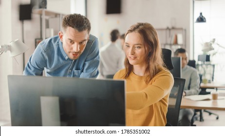 Female Specialist Works On Desktop Computer, Project Manager Stands Beside And Gives Advice On Optimizing Workflow For Customer Experience Management. Modern Office With Diverse Team Of Professionals