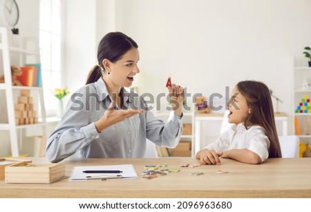 Female speech therapist working on dyslexic kid's problems and impediments. Happy child together with speech language pathologist sitting at a desk in modern office, playing fun game, learning letters