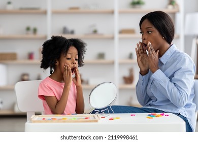 Female speech-language pathologist young black woman exercising with adorable little girl with bushy hair, working on word sonds, kid looking at mirror and touching her mouth, clinic interior