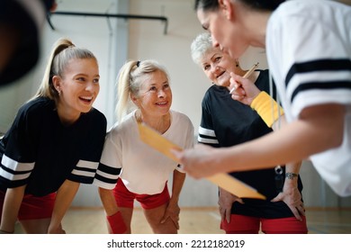 Female Sport Coach With Clipboard Discussing Tactics With Young And Old Women Team Training For Match In Gym.