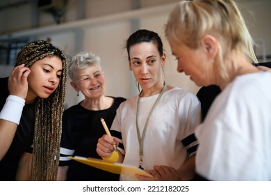 Female Sport Coach With Clipboard Discussing Tactics With Young And Old Women Team Training For Match In Gym.