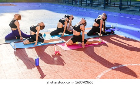 Female Sports Team Of Attractive Slender Girls Doing Yoga Exercises On Special Soft Mats On The Outdoor Stadium In The Urban Park At Sunrise. Healthcare And Active, Sports Lifestyle Concept.