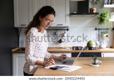 Female standing in home kitchen doing accounting work from home calculates company financial statement, use calculator fill data results on laptop application, housewife manage family budget concept