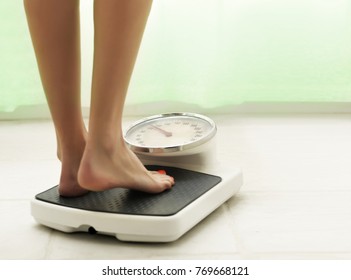 Female standing on the weight scale in the white bathroom floor with green curtain in background