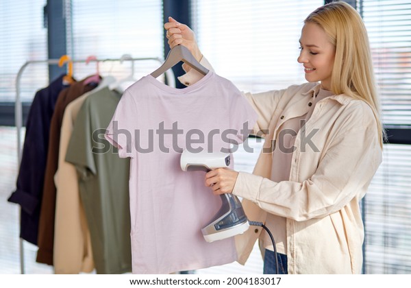 Female steams clothes on hanger. Woman with\
steamer cleaning clothes side view, copy space. Blonde caucasian\
lady using electric moder steamer, dressed casually, at home. Smart\
house concept