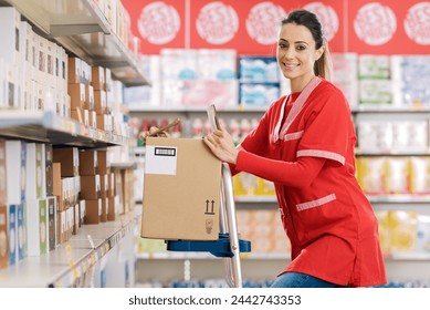 Female stock clerk working at the supermarket, she is standing on a ladder and smiling - Powered by Shutterstock