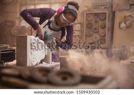 Female stonemason cutting a piece of white marble in a workshop, craftswoman grinding a sculpture with angle grinder, dust and debris flying, women doing hard work concept, stonemasonry and stonecraft