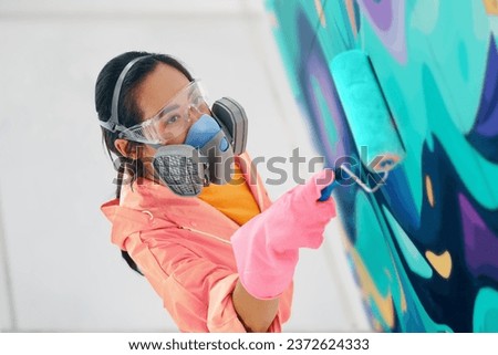 Female street artist in respirator mask painting colorful graffiti with paint roller on wall Modern art, urban concept.