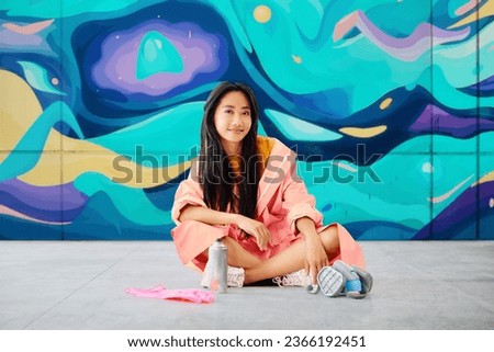 Female street artist rest sitting on the ground near the wall with her colorful paintings looking to camera. Street art concept